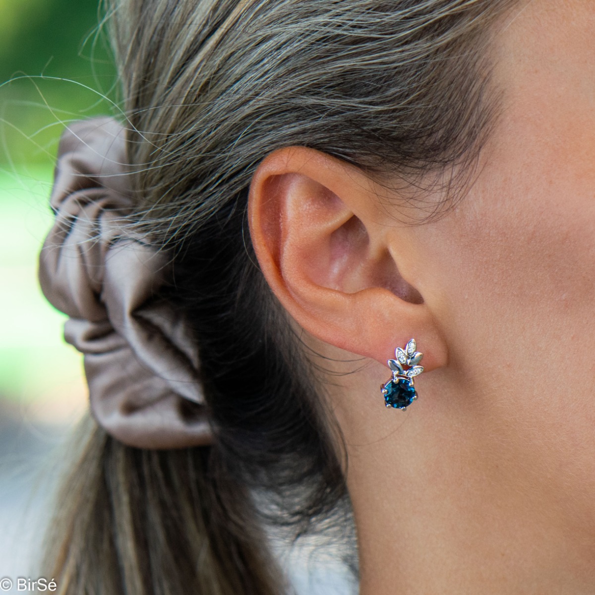 Delicate ladies' earrings with English clasp and an exclusive design combining a radiant natural London topaz surrounded by cubic zirconia petals and crafted entirely in rhodium silver. They can be combined with a pendant and a ring of the same model.