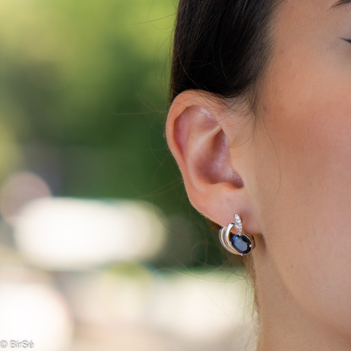 Sophisticated women's earrings with a non-standard and modern design, made entirely of beautiful rhodium-plated silver, complemented by a radiant natural sapphire stone and delicate zircons. In an exclusive sterling silver pendant and ring set.